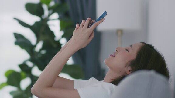 年轻的亚洲女子坐在沙发上用智能手机和她的朋友聊天
