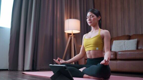 宁静的亚洲女性坐在地板上训练瑜伽在线在家练习瑜伽冥想和做呼吸练习吸气和呼气同时保持她的眼睛闭着