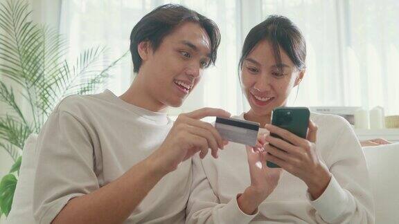 年轻的亚洲夫妇坐在家里客厅的沙发上一起玩智能手机购物使用信用卡在线支付生活方式花时间在家里