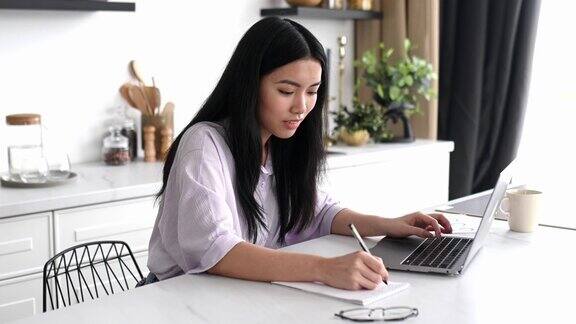 积极、漂亮、聪明的中国黑发女孩设计师、自由职业者或学生穿着衬衫坐在家里的厨房里拿着笔记本电脑远程工作或学习浏览互联网做笔记友好的微笑