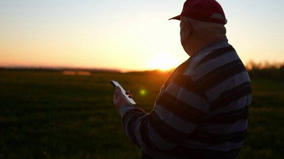 老农民拿着智能手机在地里干活从事智慧农业的资深农民拖拉机在后面行驶