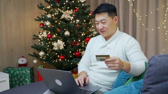 亚洲男子用笔记本电脑和信用卡在网上购物坐在家里的沙发上新年圣诞树寒假