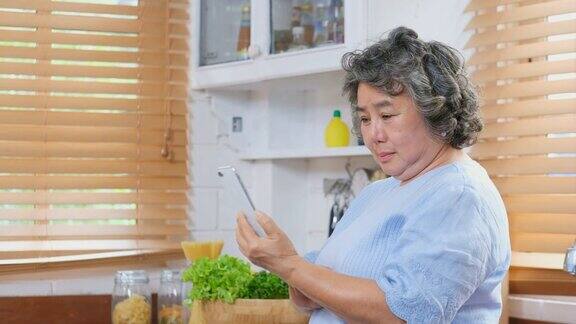 高级亚洲女性使用智能手机和在厨房喝咖啡高级人员和技术