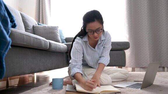 一位年轻的亚洲女商人正在笔记本电脑上寻找信息她在笔记本上做笔记她坐在家里客厅的地毯上商业女性和工作理念
