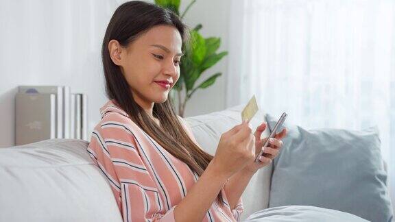 亚洲美女喜欢在家里的客厅网上购物漂亮的女孩坐在沙发上用手机在网上商店购买商品后拿着信用卡进行网上银行支付