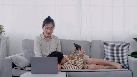 4K亚裔单亲妈妈躺在膝盖上玩平板电脑亚裔单亲妈妈坐在沙发上玩笔记本电脑男孩玩得很开心男孩躺在最舒服的位置玩