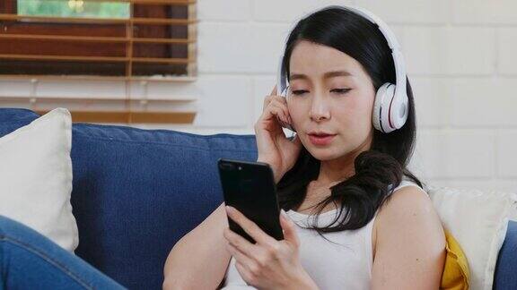 年轻的亚洲女子戴着耳机一边听着智能手机里的音乐一边坐在家里的沙发上livibg房间背景下享受着人们的生活方式