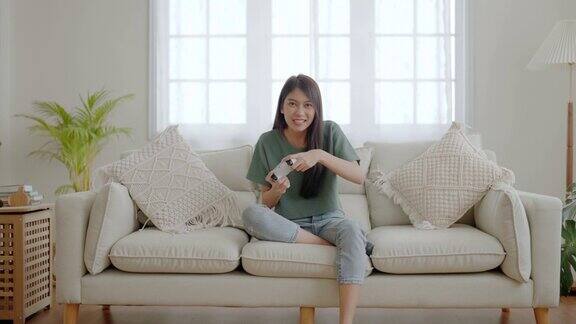 美丽的年轻亚洲女子坐在沙发上拿着操纵杆玩在线视频游戏在家里的客厅