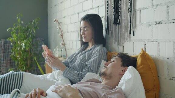 亚洲女性使用智能手机触屏微笑而混血男子在家里的床上睡觉