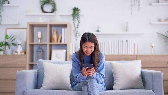 快乐的年轻亚洲女人在家里舒适的沙发上放松在智能手机上输入聊天信息微笑的女孩用手机聊天在浏览器上搜索信息无线互联网网上购物