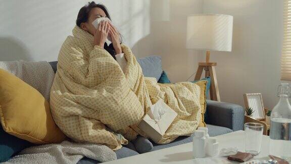 生病的年轻亚洲妇女擤鼻涕和打喷嚏用纸巾坐在毯子下的沙发在家里的客厅