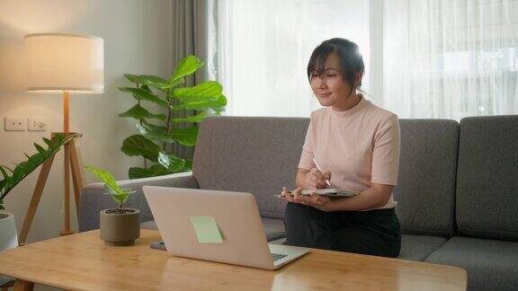 女人用笔和平板电脑看着笔记本电脑坐在家里现代客厅的沙发上