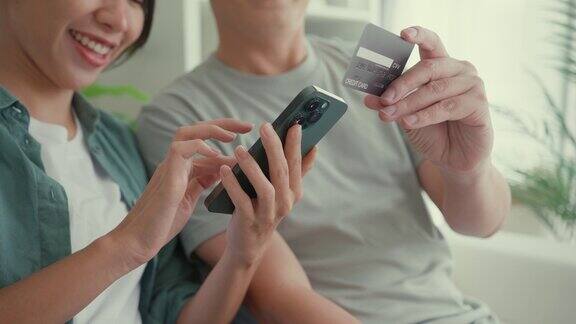 幸福的亚洲夫妇的特写他们一起坐在家里客厅的沙发上玩智能手机购物使用信用卡在线支付生活方式花时间在家里