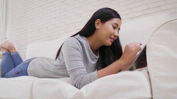 一名年轻的亚洲女性躺在家里客厅的沙发上使用数码平板电脑