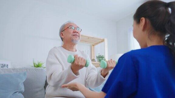 亚洲老年男子在护士的支持下做物理治疗老年男性患者坐在客厅的沙发上在养老院的治疗师医生的指导下进行哑铃锻炼