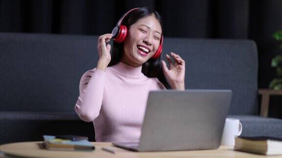 快乐的年轻亚洲女人戴着耳机在笔记本电脑上听音乐在家里跳舞