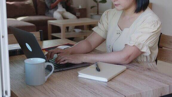 近距离的亚洲女学生打字笔记本电脑键盘而在线学习在家里的客厅她的朋友坐在旁边的沙发上