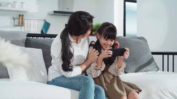 亚洲家庭母亲和女儿坐在沙发上一起在客厅玩智能手机游戏幸福和微笑快乐活动科技生活方式手机使用概念