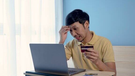 4K50fps帅气的亚洲男孩穿着黄色衬衫看着他的信用卡在他的笔记本电脑前他的表情很严肃他在网上购物网上购物