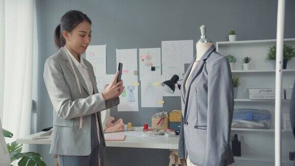 愉悦的亚洲女商人时装设计师带着套装系列坐在桌子上用智能手机拍照在工作室工作场所向客户确认采购订单启动业务
