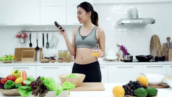 周末早上年轻的亚洲女子在厨房里使用智能手机喝着橙汁健康饮食和健康生活方式
