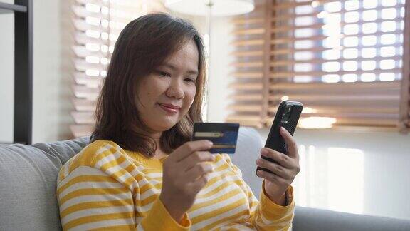 一个快乐的女人坐在沙发上用手机和信用卡在网上购物