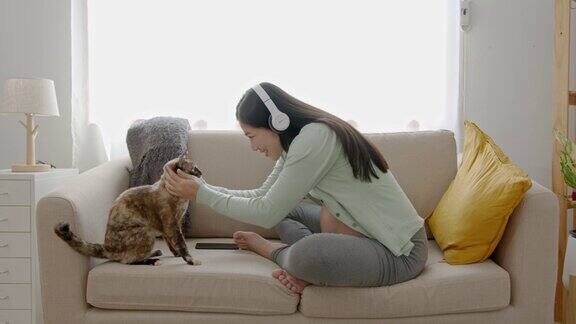 亚洲孕妇玩猫和听音乐的耳机坐在家里的沙发上怀孕产妇准备和预期