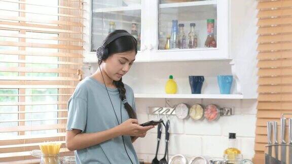 年轻美丽的亚洲女人戴着耳机和使用智能手机而站在厨房