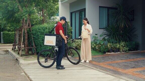 年轻的亚洲快递员骑着自行车穿着红色制服携带箱子敲门回家网上购物纸袋女人在门前女士扫描二维码支付在家里的智能手机