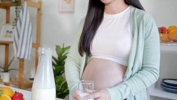 年轻的亚洲孕妇站在厨房房间拿着一杯新鲜牛奶在怀孕期间吃东西以保持身体健康直到分娩