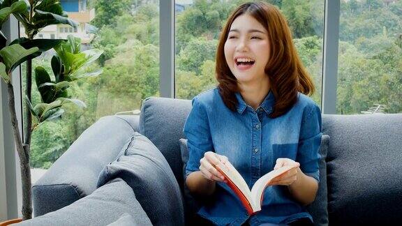 年轻的亚洲女人放松看书和在家里的沙发上笑