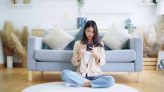 快乐的年轻亚洲女子放松在舒适的沙发上在家里发短信在手机上微笑的女孩使用手机网上聊天信息网上购物从家里