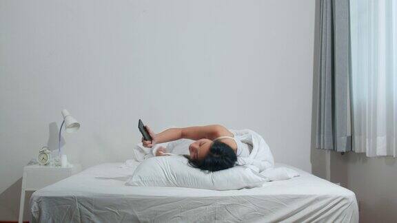 年轻的亚洲女人使用智能手机查看社交媒体早上醒来后躺在床上开心地微笑美丽迷人的西班牙女孩微笑放松在卧室在家里