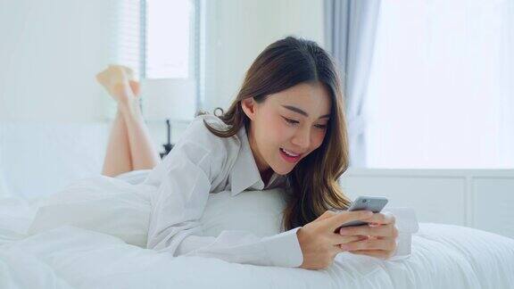 亚洲美女早上在家里的床上用手机聊天快乐随意的漂亮女性躺在卧室享受假日周末在家里用智能手机和科技交流