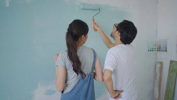年轻的亚洲夫妇在他们的新公寓装修期间一边用滚轴修理和刷蓝色油漆的墙壁一边跳舞