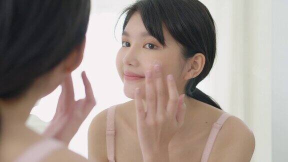 美丽的年轻亚洲女人微笑看着镜子检查脸和皮肤护理霜和化妆品为年轻化和卫生美丽的女孩快乐清洁面部与乳液和健康
