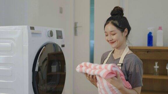 闻一闻笑一笑年轻的亚洲女人在家里用洗衣机洗衣服快乐的女孩和一个家庭清洁工闻到毛巾或衣服从洗涤幸福和家务