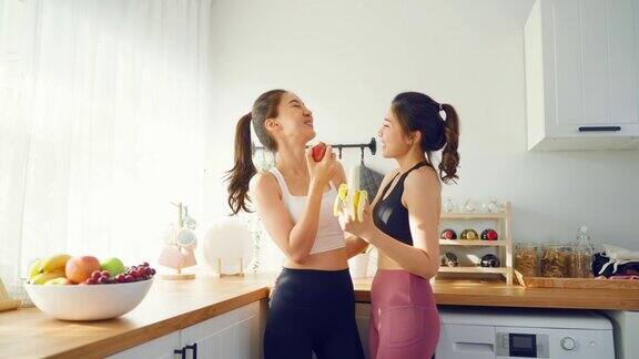 亚洲活跃的两个女人姐妹穿着运动服在厨房吃苹果年轻美丽的女孩妹妹心情愉快享受吃水果健康的食物来饮食和减肥的保健在家里