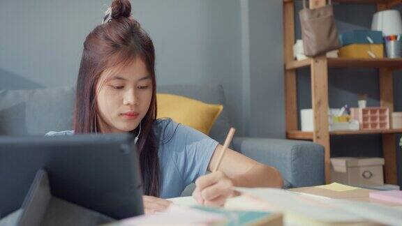 年轻的亚洲女孩青少年戴着休闲耳机使用数码平板电脑在线学习写演讲笔记本在客厅在家里隔离教育在线电子学习冠状病毒大流行概念