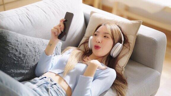 快乐的年轻亚洲女性在家里放松坐在沙发上拿着手机的女性微笑着女孩用视频打电话给朋友听音乐