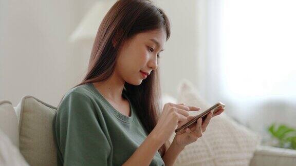 快乐的年轻亚洲女人放松在舒适的沙发在家里用智能手机发短信微笑的女孩用手机聊天浏览无线网络小工具从家里在线购物
