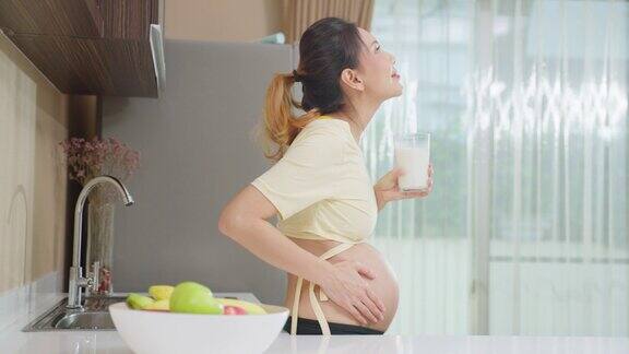 亚洲美女怀孕站在家里喝牛奶迷人的孕妇站在子宫里抚摸宝宝喝鲜奶健康饮料在厨房里感受幸福