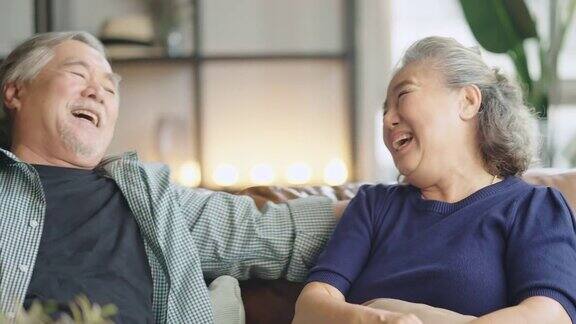 亚洲老老人退休夫妇享受在沙发上谈笑风生在家里开心地笑着亚洲老成年人居家隔离期
