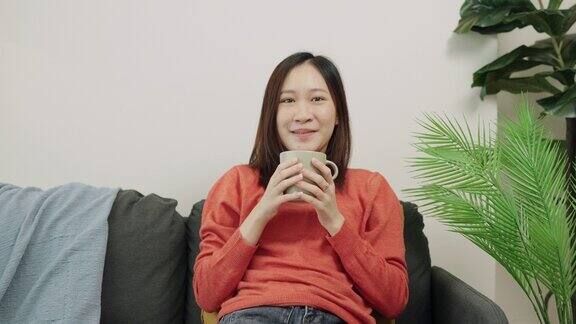 亚洲放松的女人坐在客厅舒适的沙发上在家里喝咖啡茶享受周末的休闲活动