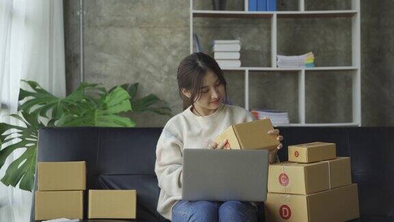 亚洲妇女坐在沙发上坐在家里的盒子和笔记本电脑从客户网上业务中小企业销售概念