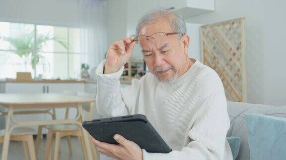亚洲魅力老人在家客厅使用平板电脑年长成熟的爷爷戴着眼镜坐在沙发上用智能手机聊天与家人交流享受退休生活
