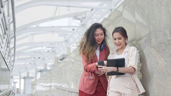 商务通勤东南亚女商人拿着咖啡杯和数码平板电脑和同事一起走在城市建筑的背景下工作