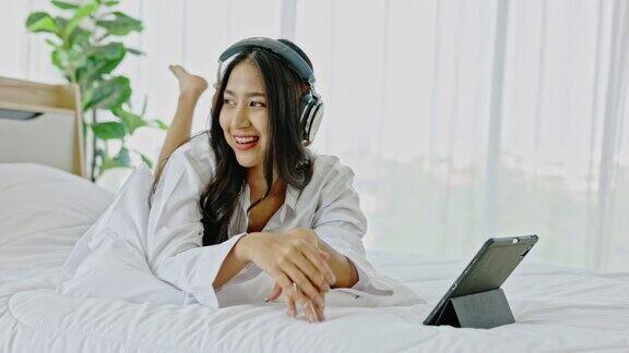 一名年轻的亚洲女性愉快地戴着耳机用笔记本电脑听在线音乐在卧室放松娱乐是生活中的一种小奢侈