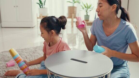 亚洲蹒跚学步的小女孩和妈妈坐在地毯上快乐的时刻玩彩色堆叠杯玩具在客厅的桌子在家里