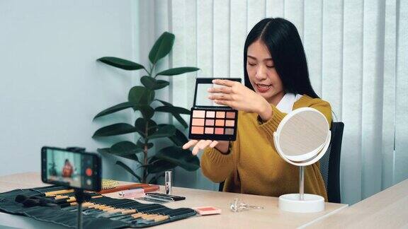 一位亚洲女性在视频博客上展示化妆品同时通过手机解释产品的使用